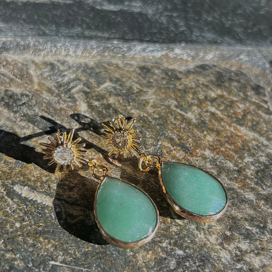A pair of gold-colored earrings featuring green aventurine stones, displayed against a natural stone background.