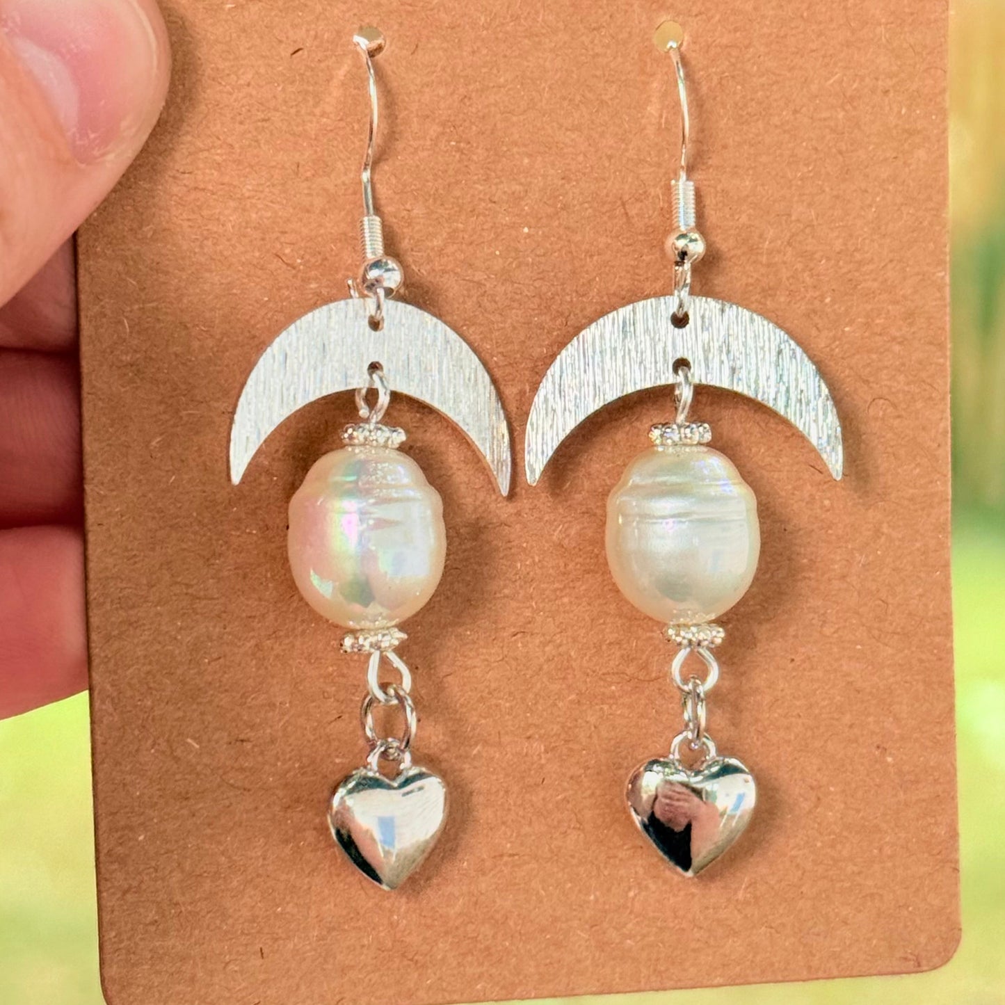 Pair of earrings with heart and moon designs on a brown card, held by a hand.