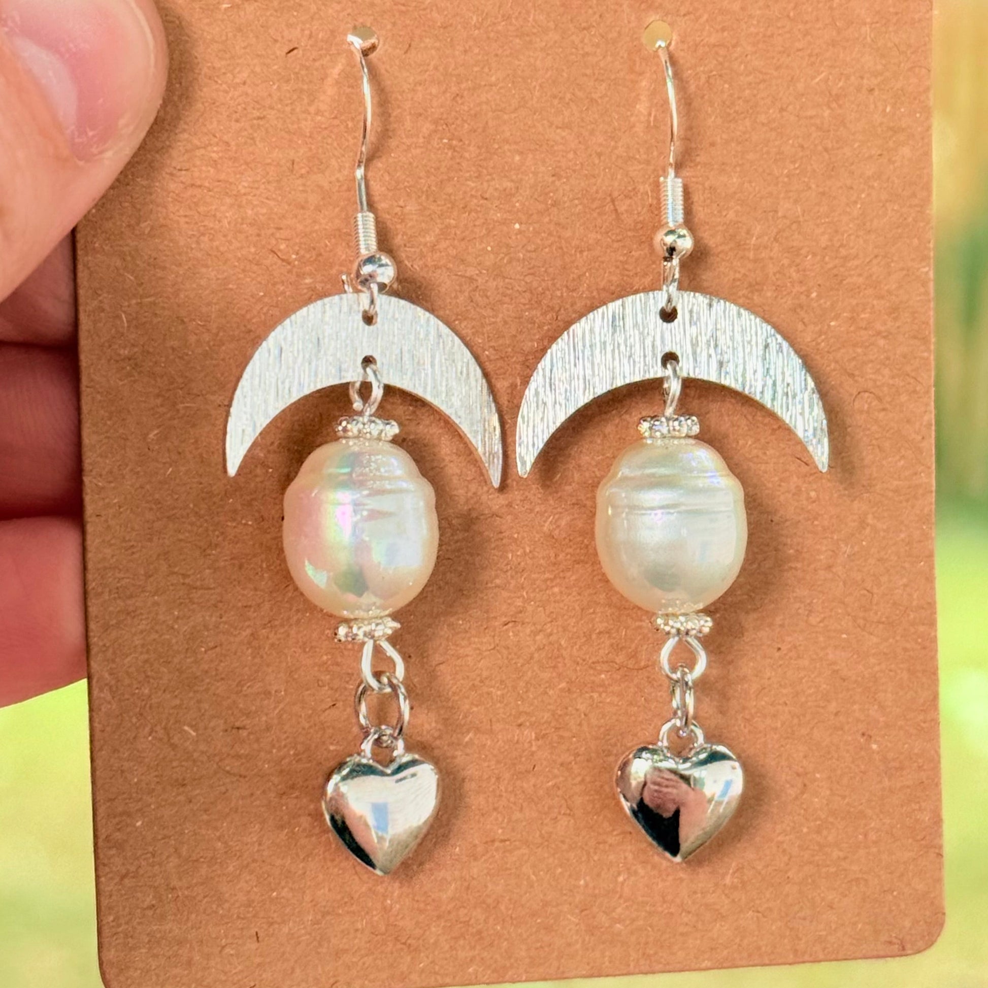 Pair of earrings with heart and moon designs on a brown card, held by a hand.