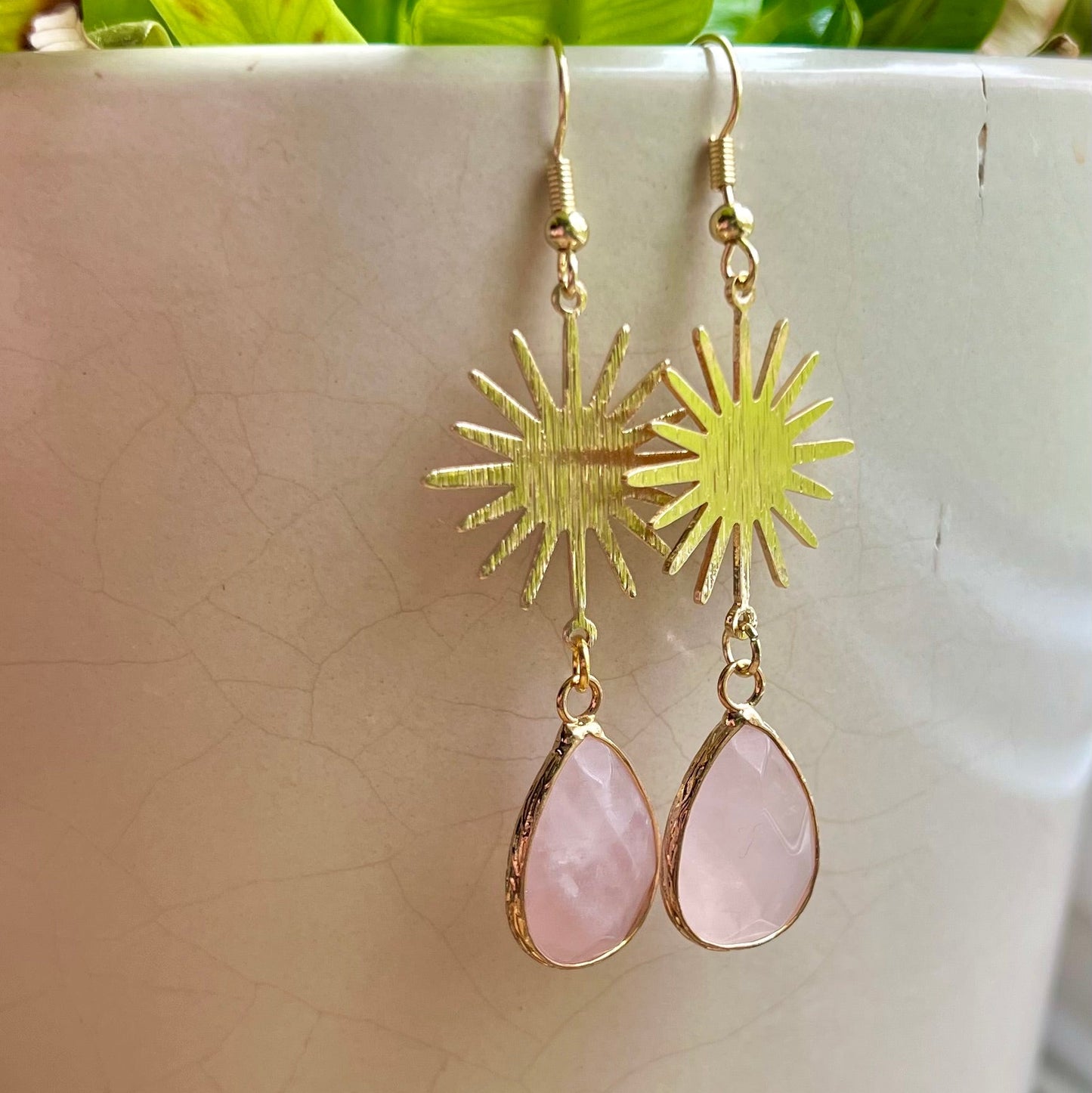A pair of gold-colored earrings with one featuring a sunburst design and the other with a teardrop-shaped pink stone, displayed against a white background with green foliage in the backdrop.