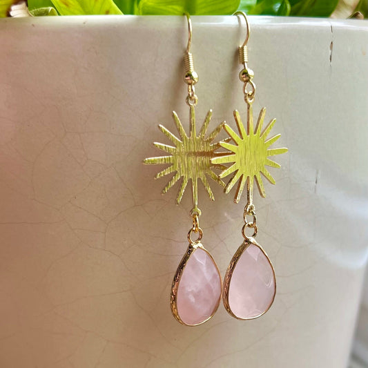 A pair of gold-colored earrings with one featuring a sunburst design and the other with a teardrop-shaped pink stone, displayed against a white background with green foliage in the backdrop.