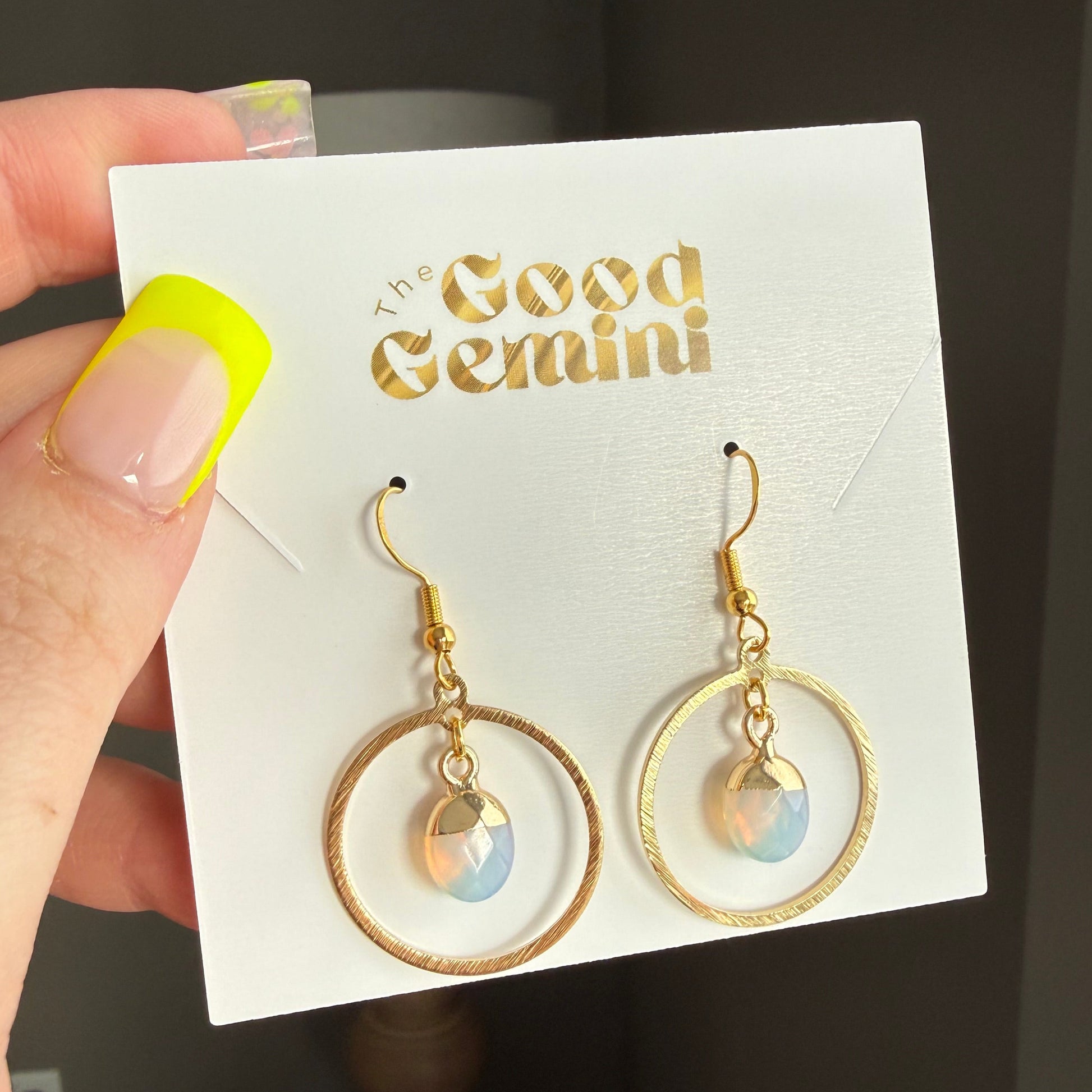 Gold earrings on a 'Good Gemini' card held by a hand with yellow nail polish.