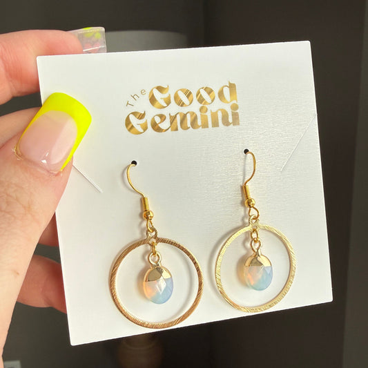 Gold earrings on a 'Good Gemini' card held by a hand with yellow nail polish.