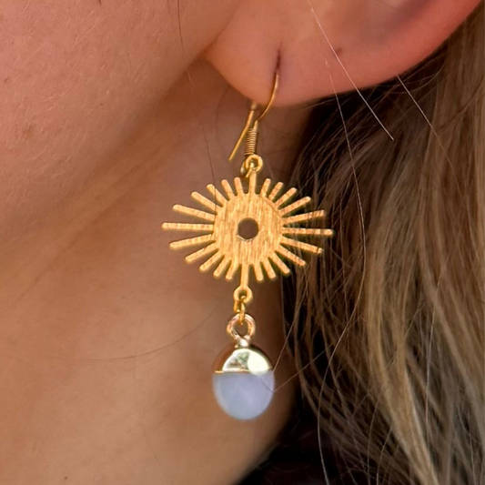 Gold sun-shaped earring with a white stone hanging from it, worn by a person.