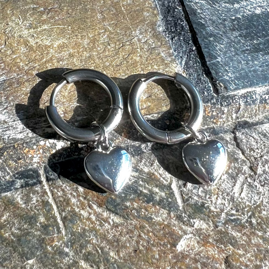 A pair of silver hoop earrings with heart charms attached, displayed on a textured surface.